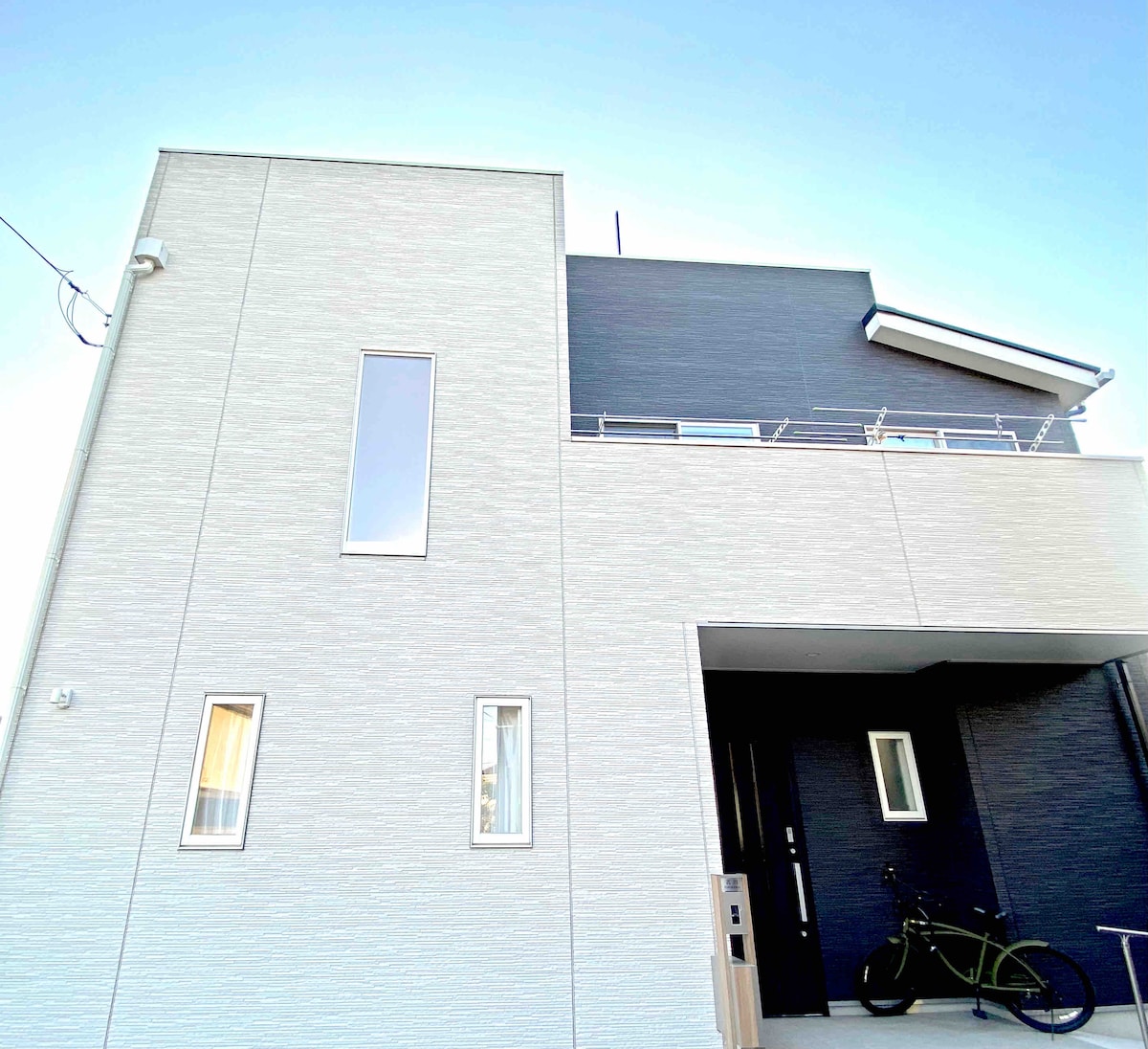 The modern exterior of the house features a combination of smooth and textured surfaces. Large windows provide natural light, and a bicycle is parked on the entrance, suggesting accessibility. The structure's clean lines contribute to a contemporary aesthetic.