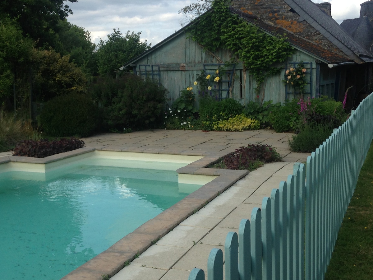 A serene swimming pool is framed by neatly arranged stone tiles, surrounded by lush green shrubs and vibrant flowers. A rustic house with weathered blue siding is visible in the background, complemented by a small garden area and a light-colored fence.