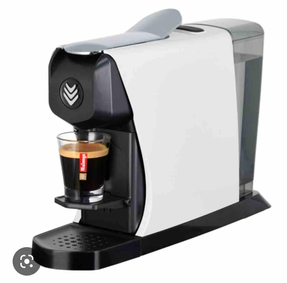 A modern coffee machine is displayed, featuring a sleek white and black design. A glass filled with freshly brewed espresso sits underneath the dispenser, showcasing the machine's functionality. The perforated drip tray allows for easy cleaning, reflecting a minimalist style.