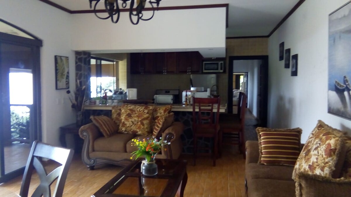 A living area features a warm color palette with a comfortable sofa and armchairs adorned with decorative pillows. A wooden table sits in the center, complemented by a bouquet of flowers. The open kitchen is visible in the background, featuring wooden cabinetry and modern appliances.