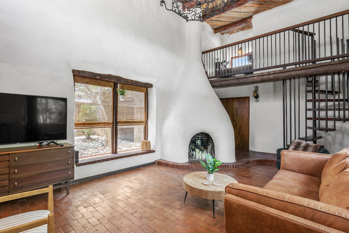A spacious living room features a prominent fireplace with a unique design, complemented by large windows allowing natural light to fill the space. A comfortable sofa and a small coffee table are arranged to create a welcoming area for relaxation. A staircase with wooden railing leads to an upper level.