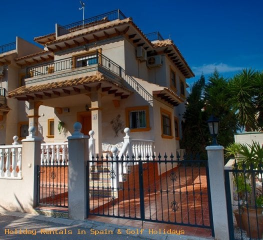 The exterior of the two-story house is shown, featuring a gated entrance with wrought iron detailing. Terracotta tiles adorn the pathway leading to the entrance, while potted plants contribute to the greenery. The facade has warm earth tones, complemented by a balcony on the upper floor.