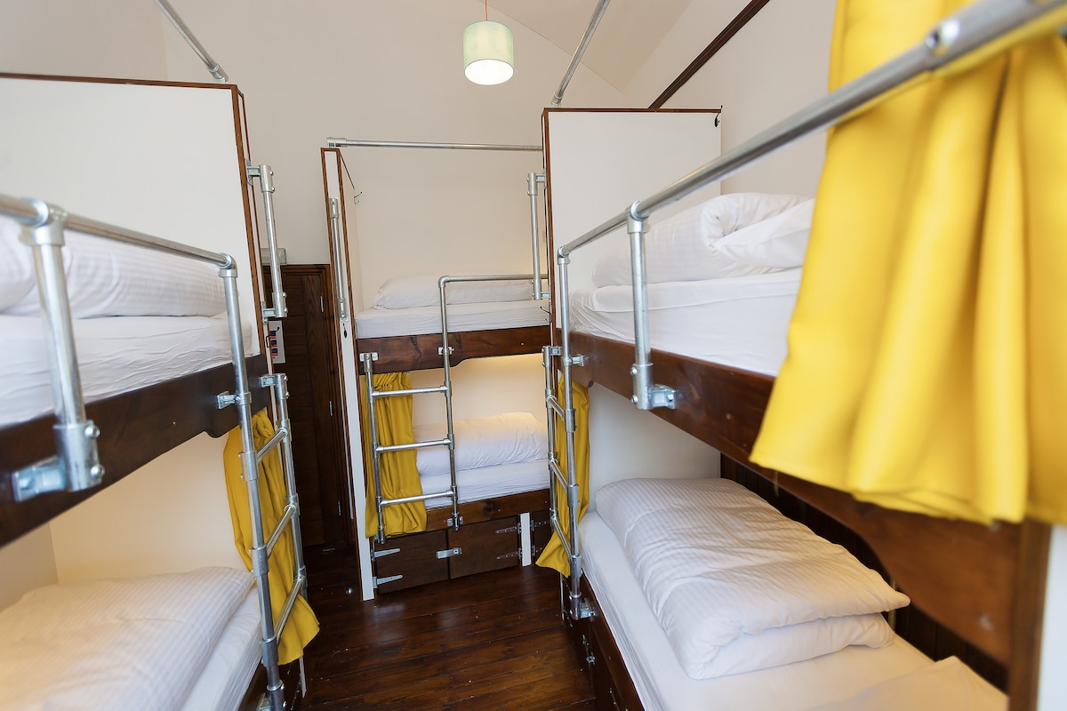 A shared room features multiple bunk beds with individual privacy curtains in vibrant yellow. Each bed includes plush bedding and a sturdy ladder provides access to the upper bunks. Natural light is present, highlighting the wooden flooring and warm tones of the space.