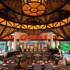 A spacious dining area is featured under a high thatched roof, adorned with warm amber lighting and vibrant orange accents. Round tables are set with white tableware, while wicker chairs offer comfort. The space is surrounded by lush greenery, creating a calming atmosphere.