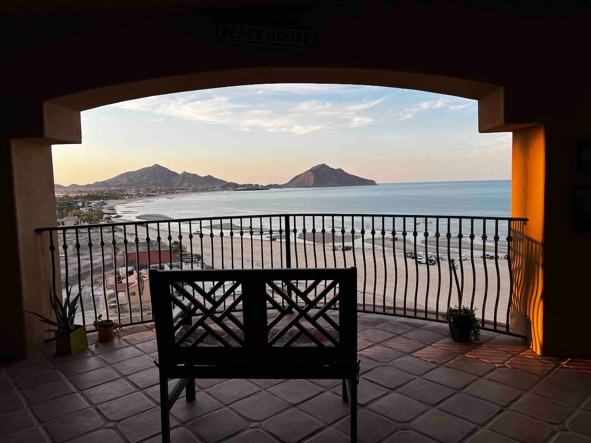 A spacious terrace view frames a scenic coastline with gentle waves lapping at the sandy shore. A silhouette of a wooden bench overlooks the beach and mountains, while planters with greenery add a touch of nature to the area. The sky exhibits warm hues from the setting sun.