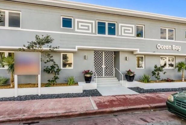 A modern building exterior is showcased, featuring a sleek grey facade and decorative Greek key details. Welcoming stairs lead to the entrance, flanked by potted plants and greenery. Large windows allow natural light to illuminate the space, contributing to an inviting appearance.