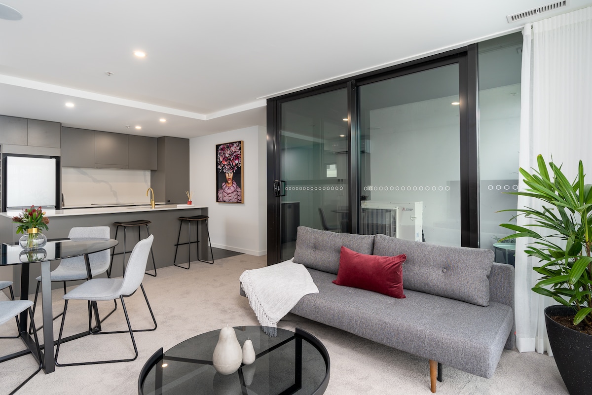 An open-concept living area is presented, featuring a stylish gray sofa with a red accent pillow. A round dining table with four chairs is positioned near the kitchen, which is equipped with modern cabinetry. Natural light fills the space through large glass sliding doors.