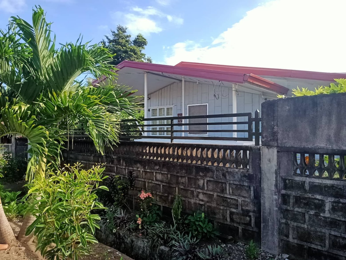 A single-story home is set behind a low stone wall, surrounded by lush greenery. The red roof contrasts with the white exterior, while large windows allow natural light to enter. Tropical plants and flowers add a touch of color to the front garden.