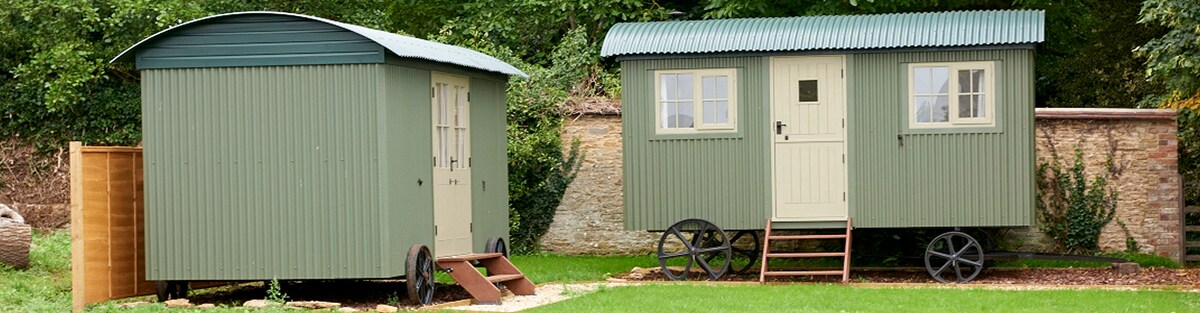 Two green, luxury shepherd's huts are visible, set in a lush garden. One hut features a small staircase leading to its entrance, while the other has a door and windows. The surrounding space offers a tranquil atmosphere, enhanced by natural greenery.
