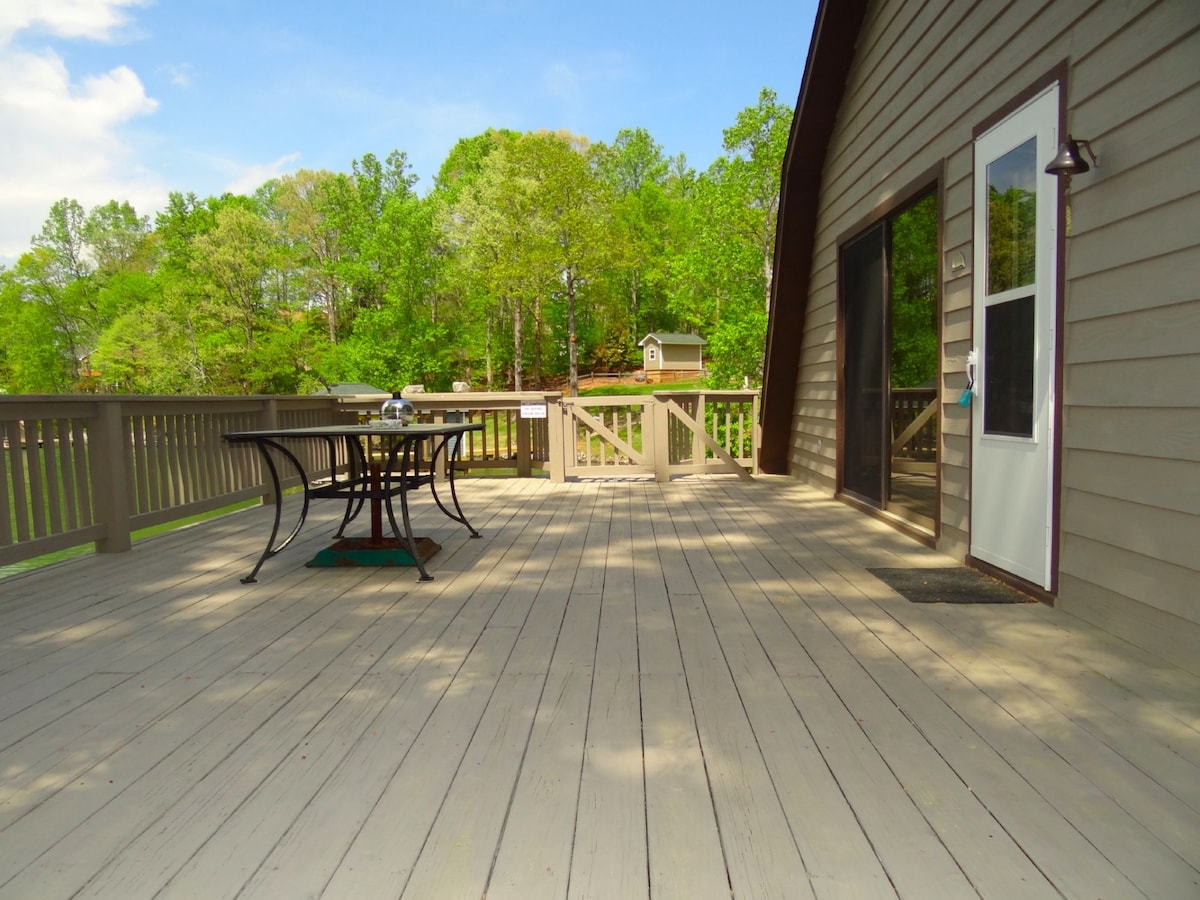 A spacious deck is featured, showcasing a dining set with a table and chairs. The wooden planks of the deck extend towards a nearby entrance. Lush greenery surrounds the area, enhancing the serene outdoor setting.