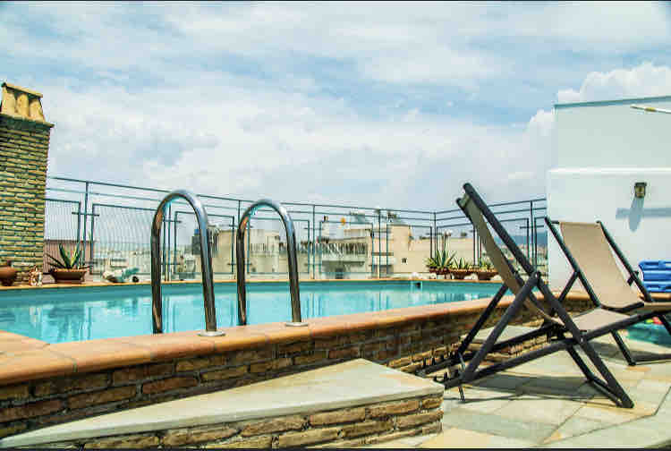 A rooftop pool is visible, surrounded by a stone deck and featuring two reclining lounge chairs. The pool reflects the sky and cityscape, with potted plants placed strategically along the perimeter. The scenery includes rooftops and distant buildings under a bright sky.