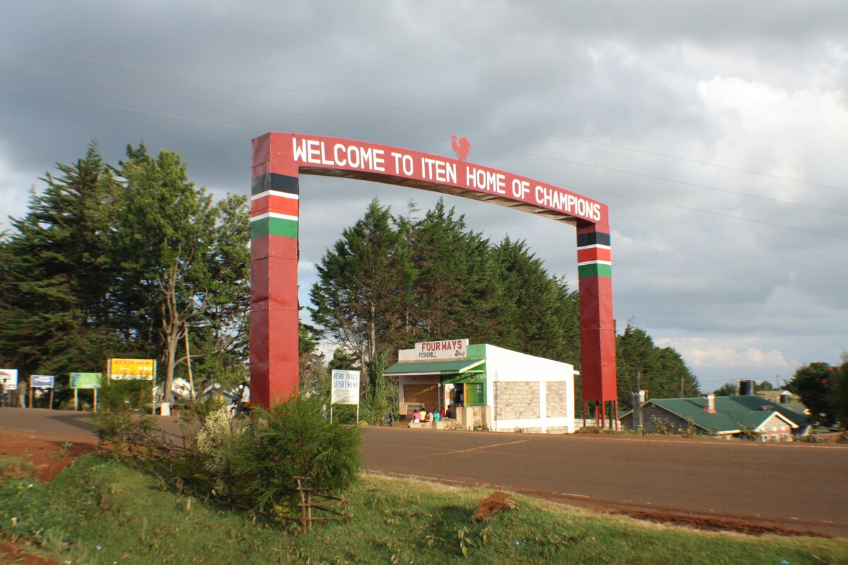 A large welcoming archway marked with the phrase 'WELCOME TO ITEN HOME OF CHAMPIONS' stands at the entrance. The structure features red, green, and black colors. Surrounding trees provide a natural backdrop, while a small building is visible nearby.