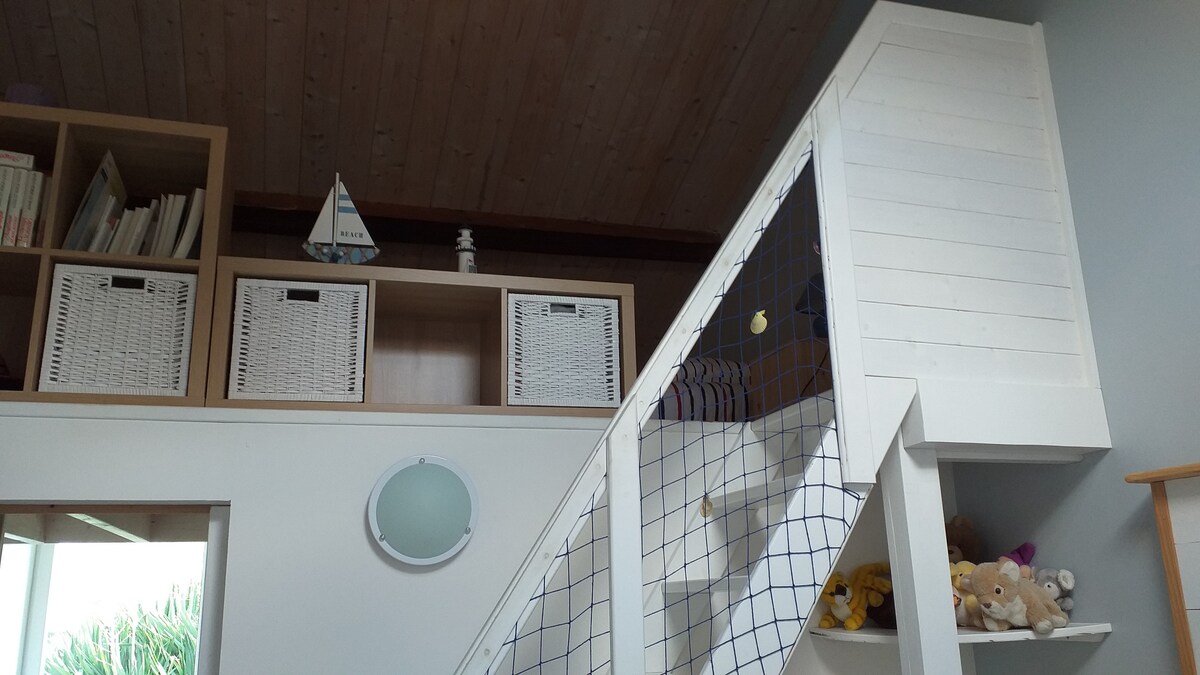 A staircase leads to a mezzanine area with wooden beams above. Storage baskets are arranged on shelving, and a small decorative sailboat adds a playful touch. A soft glow is provided by a round wall light, contributing to the bright interior.