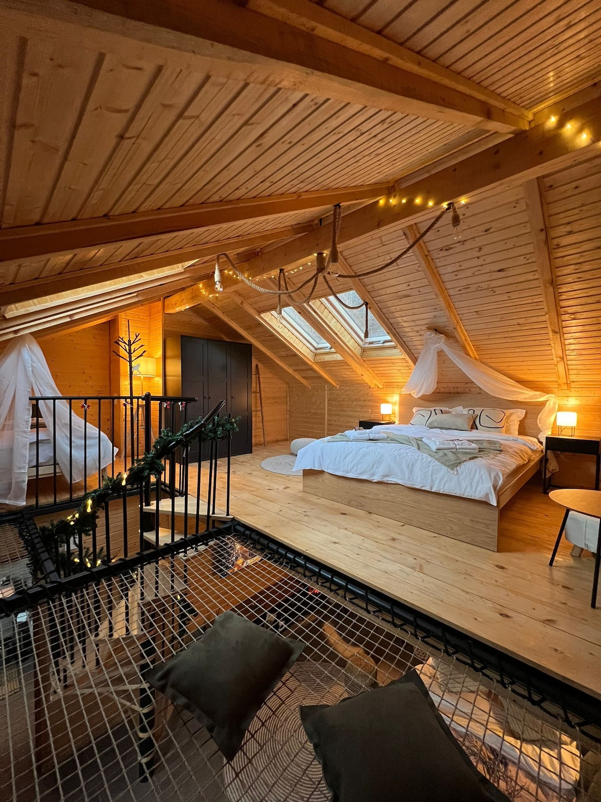 An airy loft space is illuminated by natural light from skylights. A spacious bed is adorned with soft linens, while seating areas are placed on a raised platform. Decorative string lights add a touch of warmth to the wooden beams and paneling overhead.