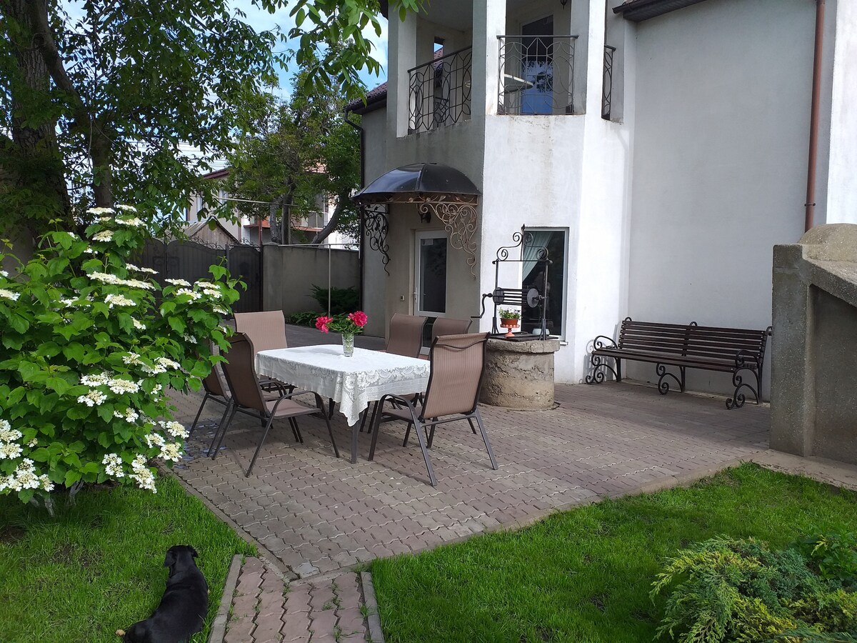 A spacious outdoor area features a dining table covered with a lace tablecloth and surrounded by several chairs. A cozy seating area with a decorative gazebo is visible, along with a grill station. Lush green grass and flowering shrubs encircle the patio.