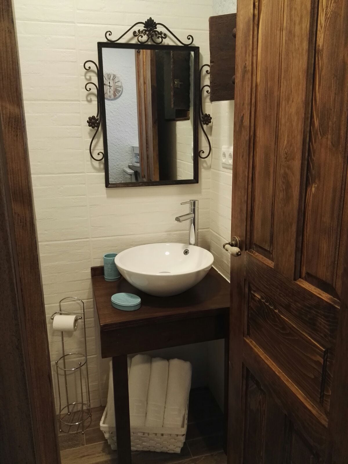 A compact bathroom area is presented, featuring a stylish round sink set atop a wooden vanity. A decorative mirror hangs above the sink. Soft towels are neatly arranged on a shelf below, while toiletries are organized beside the sink for convenience.