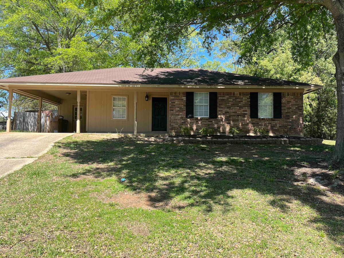A single-story home is set amidst greenery, featuring a brick facade and a welcoming front door. Large windows provide natural light to the interior. A carport is visible on the left, with an expansive lawn that enhances the tranquil environment.