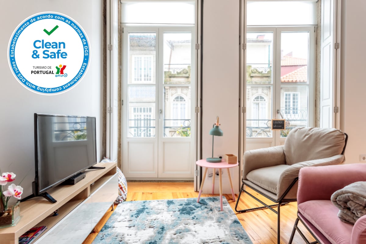 A bright living area features a spacious television stand with a TV, complemented by a plush armchair and a small side table. Light filters in through large windows, providing views of the outdoors. A decorative rug adds a touch of color to the wooden floor.