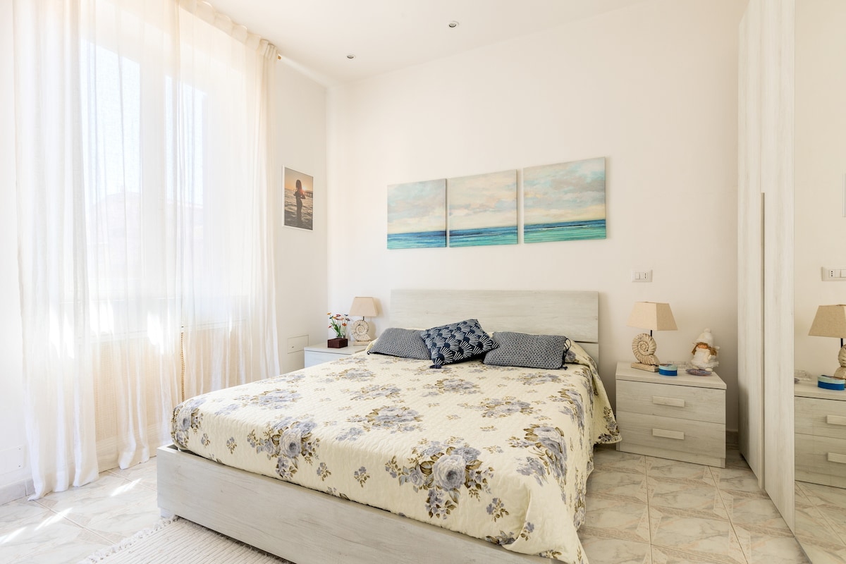 A spacious bedroom features a large bed with a floral-patterned bedspread. Two bedside lamps sit on nightstands, and a soft blue decorative pillow complements the bed. Light filters in through sheer curtains, illuminating the room. Three coastal-themed canvases adorn the wall above the bed.