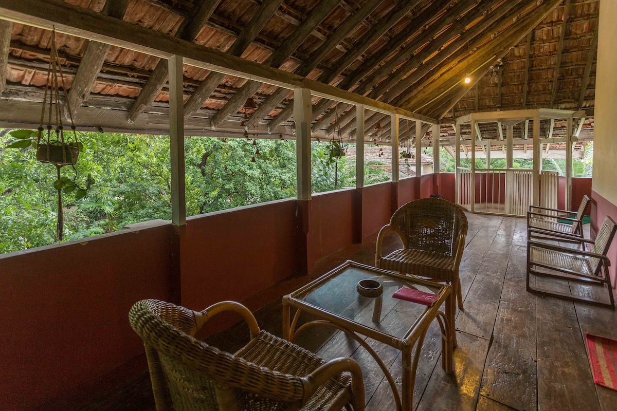 A shared balcony area features traditional wooden beams and a tiled roof. Wicker chairs and a glass-top table occupy the space, enhancing a relaxed atmosphere. Lush greenery is visible through the railing, while wooden flooring adds a rustic charm to the setting.