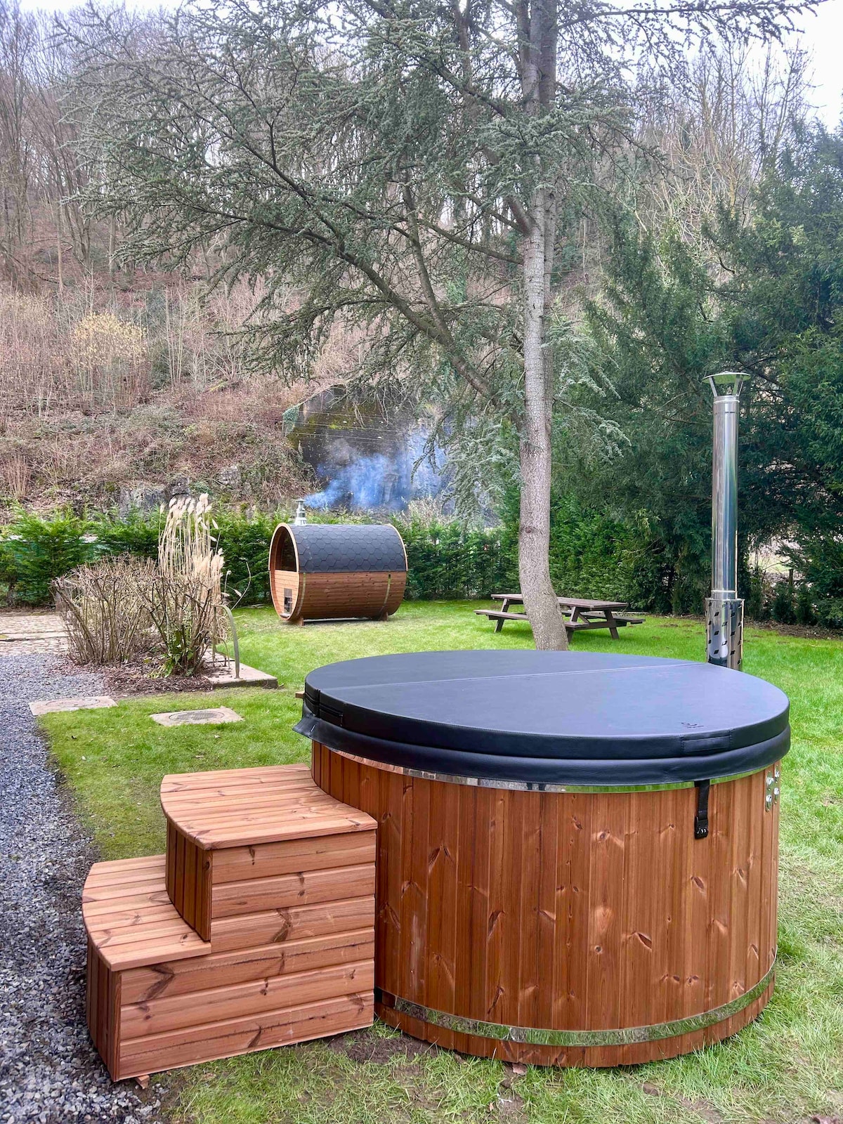 A wooden hot tub is positioned in a grassy area, featuring a black cover and wooden steps for easy access. Nearby, a cylindrical sauna is visible among greenery, with smoke gently rising from its chimney. A serene natural backdrop enhances the outdoor relaxation space.