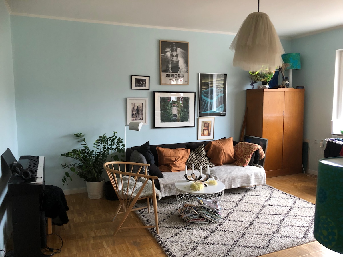 A light-filled living area features a comfortable sofa adorned with various cushions. Wall art is displayed alongside a vibrant plant. A unique light fixture hangs above a coffee table, surrounded by a woolen rug and wooden furniture, creating an inviting setting.