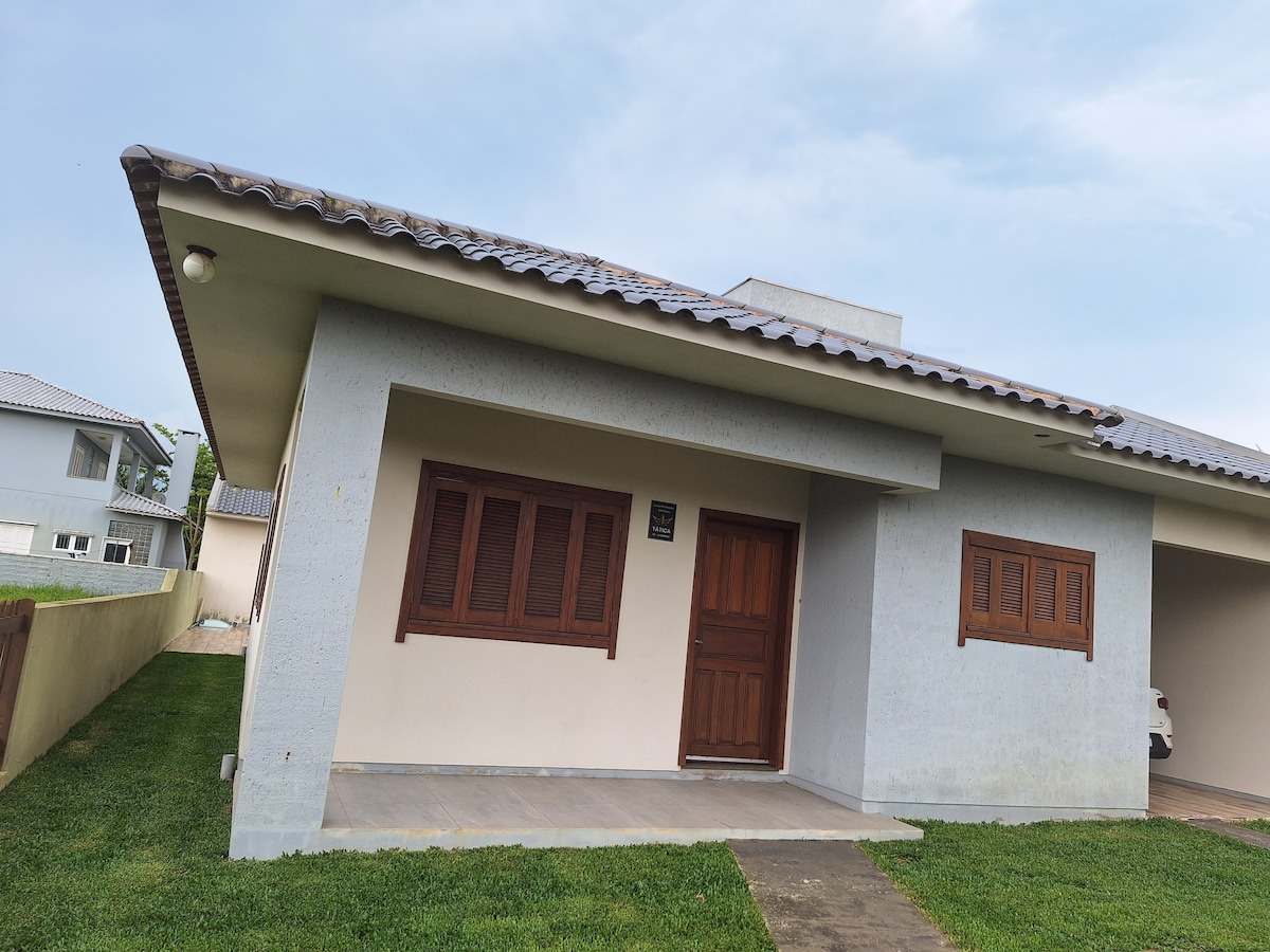 The exterior of the house is depicted, showcasing a minimalist design with a light-colored facade and a sloped roof. Wooden shutters frame the windows, and a wooden front door adds warmth. A grassy area surrounds the entrance, leading to a paved pathway.