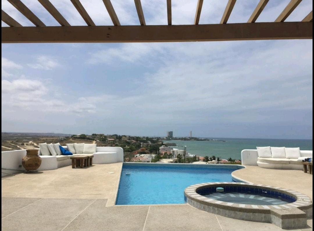 A serene outdoor space is highlighted by a sparkling pool and a circular hot tub. Comfortable seating is arranged under a wooden pergola, providing shade. The expansive view of the coastline and distant buildings adds to the peaceful atmosphere.