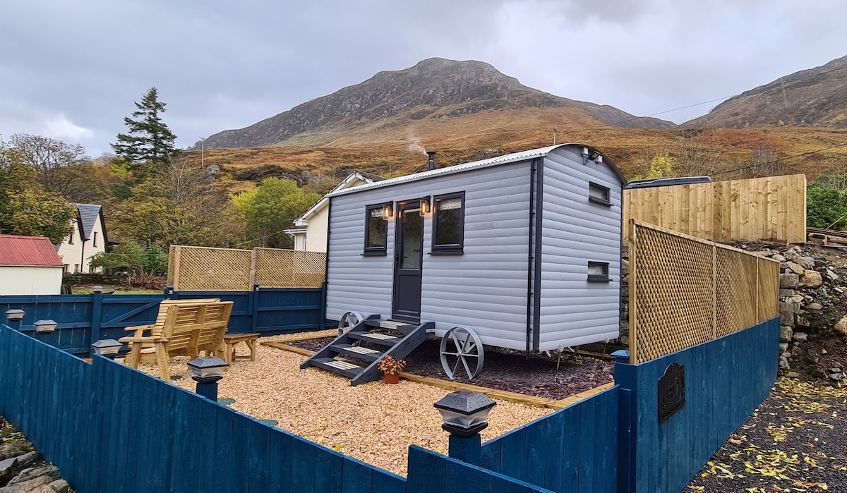 A handcrafted shepherd's hut is set against a mountain backdrop, enclosed by a wooden fence. The exterior features a light grey color with decorative wheels. A wooden deck leads to a gravel area with a picnic table and benches, inviting relaxation in the serene environment.