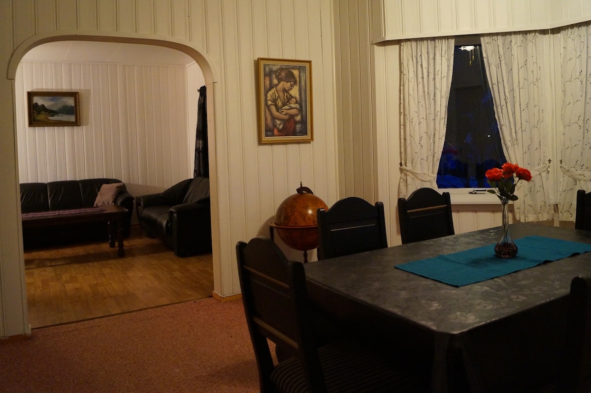 A dining area features a dark table with a blue cloth and a floral centerpiece. Six black chairs are positioned around the table. An archway leads to a cozy living space with two sofas and a painting hanging on the wall, where natural light filters through a curtained window.