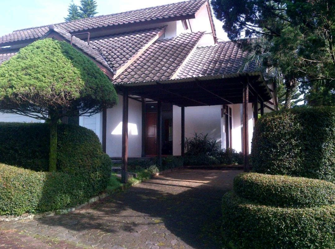 The entrance of the villa is framed by neatly trimmed shrubbery and lush greenery. A covered porch leads to the main door, while a stone pathway is visible beneath the shaded canopy. The architecture features a tiled roof and a combination of white walls and wooden accents.