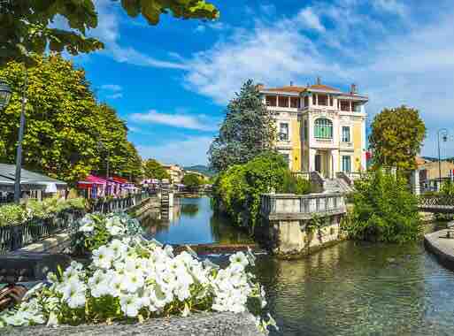 A scenic view of a charming building situated alongside a calm canal, flanked by lush greenery and blooming white flowers. Outdoor seating with colorful umbrellas is visible along the waterway, contributing to a serene atmosphere under a clear blue sky.