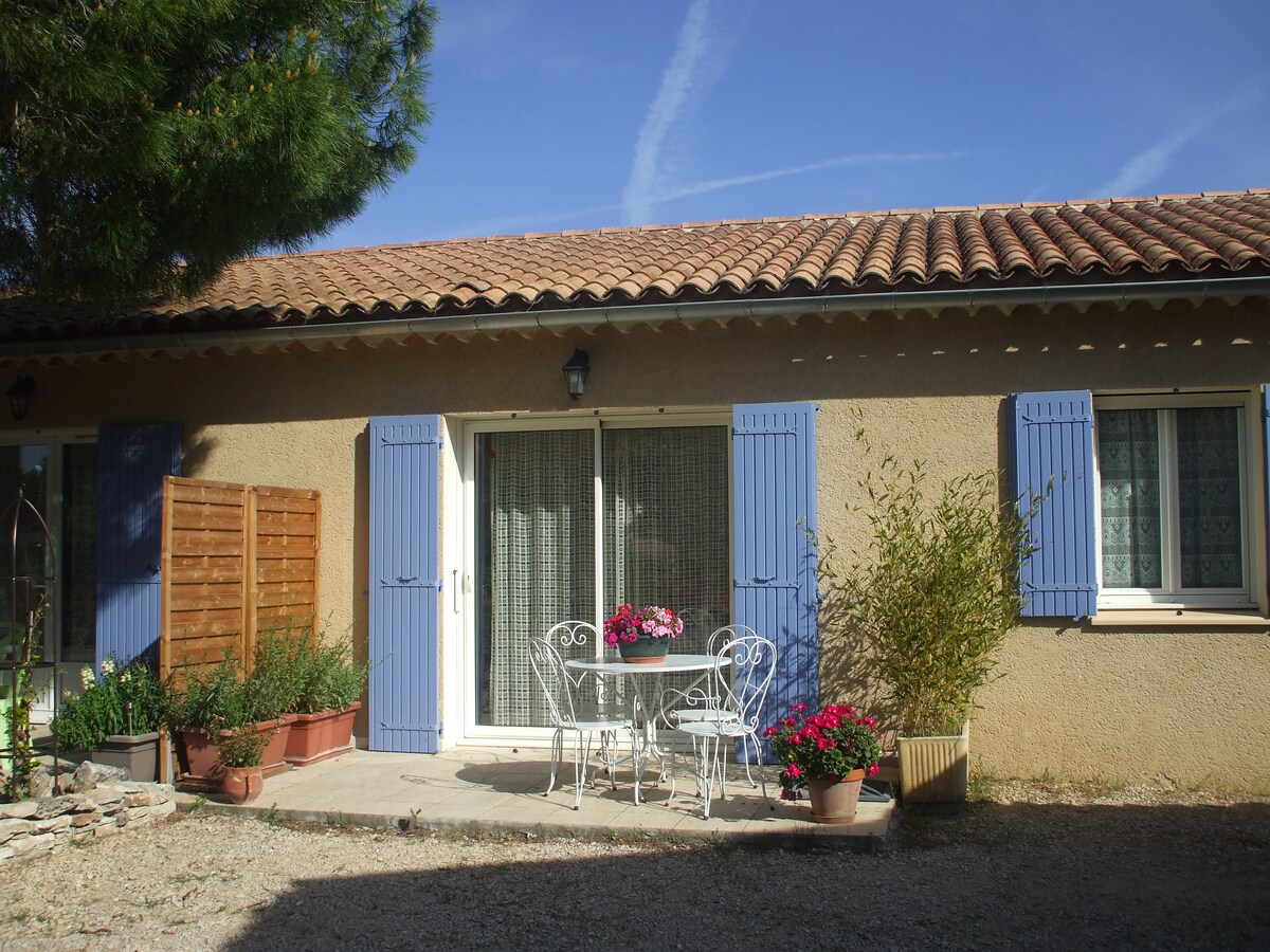 The exterior of the residence is showcased, featuring a tiled roof and light-colored walls. A small patio area is visible, furnished with a table and two chairs, adorned with potted flowers. Blue shutters frame the large sliding door, which leads to the indoor space.