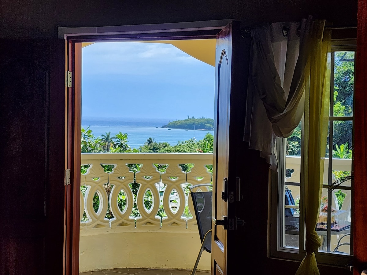 A view of the Caribbean Sea is framed by an open door, leading to a balcony with decorative railing. Lush greenery and distant land features are visible outside, enhancing the serene atmosphere of the retreat.