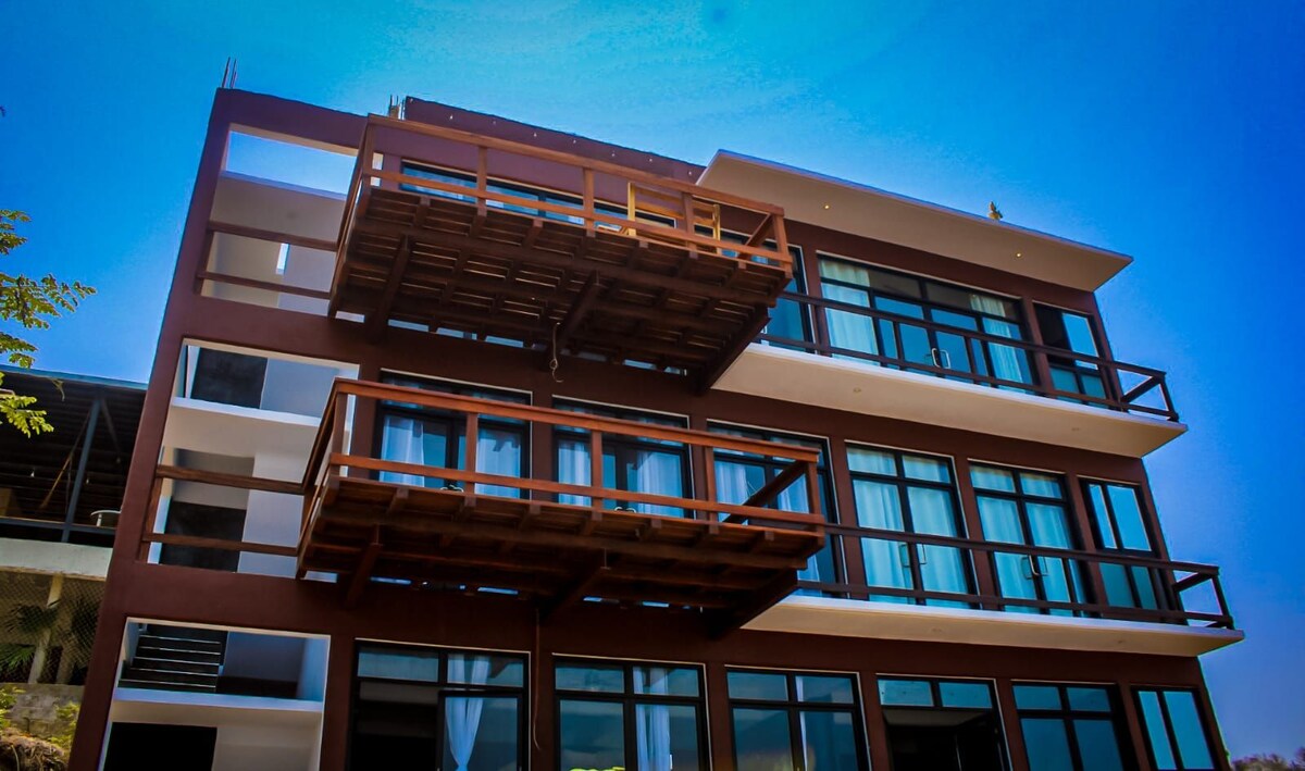 A modern three-story building features large glass windows and multiple wooden balconies. The structure is set against a clear blue sky, showcasing a clean and contemporary architectural design.