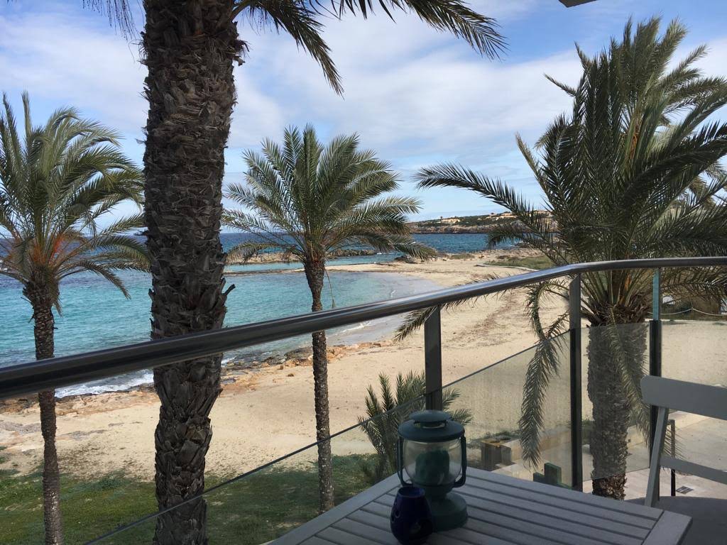 A balcony view showcases swaying palm trees overlooking a sandy beach and turquoise waters. The horizon is dotted with gentle waves, while a lantern rests on a table, completing the serene outdoor atmosphere.