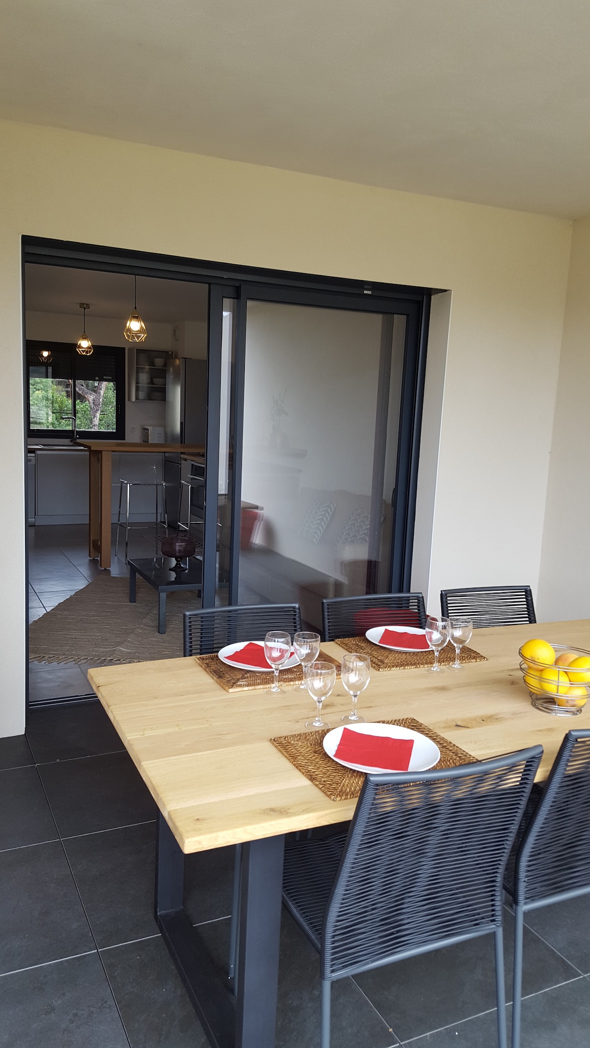 A dining table with a natural wood finish is set for a meal, featuring clear glassware and red napkins. The sliding glass doors provide a view into the kitchen area. A bowl of lemons adds a pop of color to the table, enhancing the inviting ambiance.