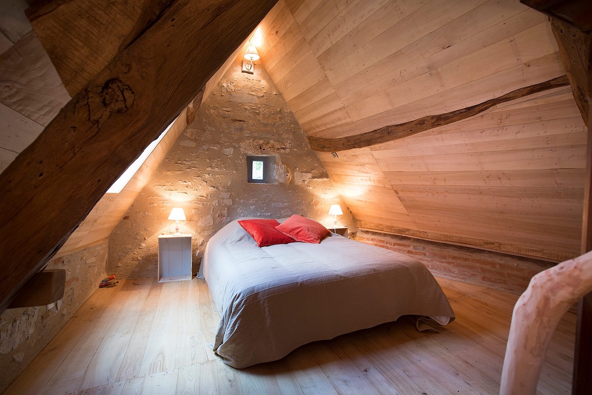 A cozy attic bedroom features sloped wooden ceilings, showcasing exposed beams and warm wooden walls. A spacious bed is adorned with neutral linens and red accent pillows. Two bedside lamps provide soft lighting, creating a serene atmosphere. A small window lets in natural light.