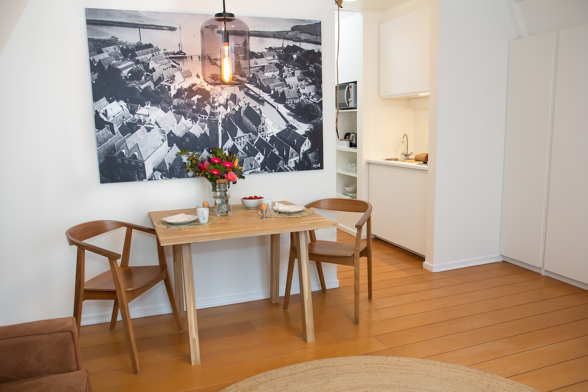 A dining area features a wooden table set for two, with a vase of flowers at the center. Two wooden chairs are placed around the table. In the background, a large black-and-white photograph captures aerial views of a town. A kitchenette is visible, providing functional amenities.