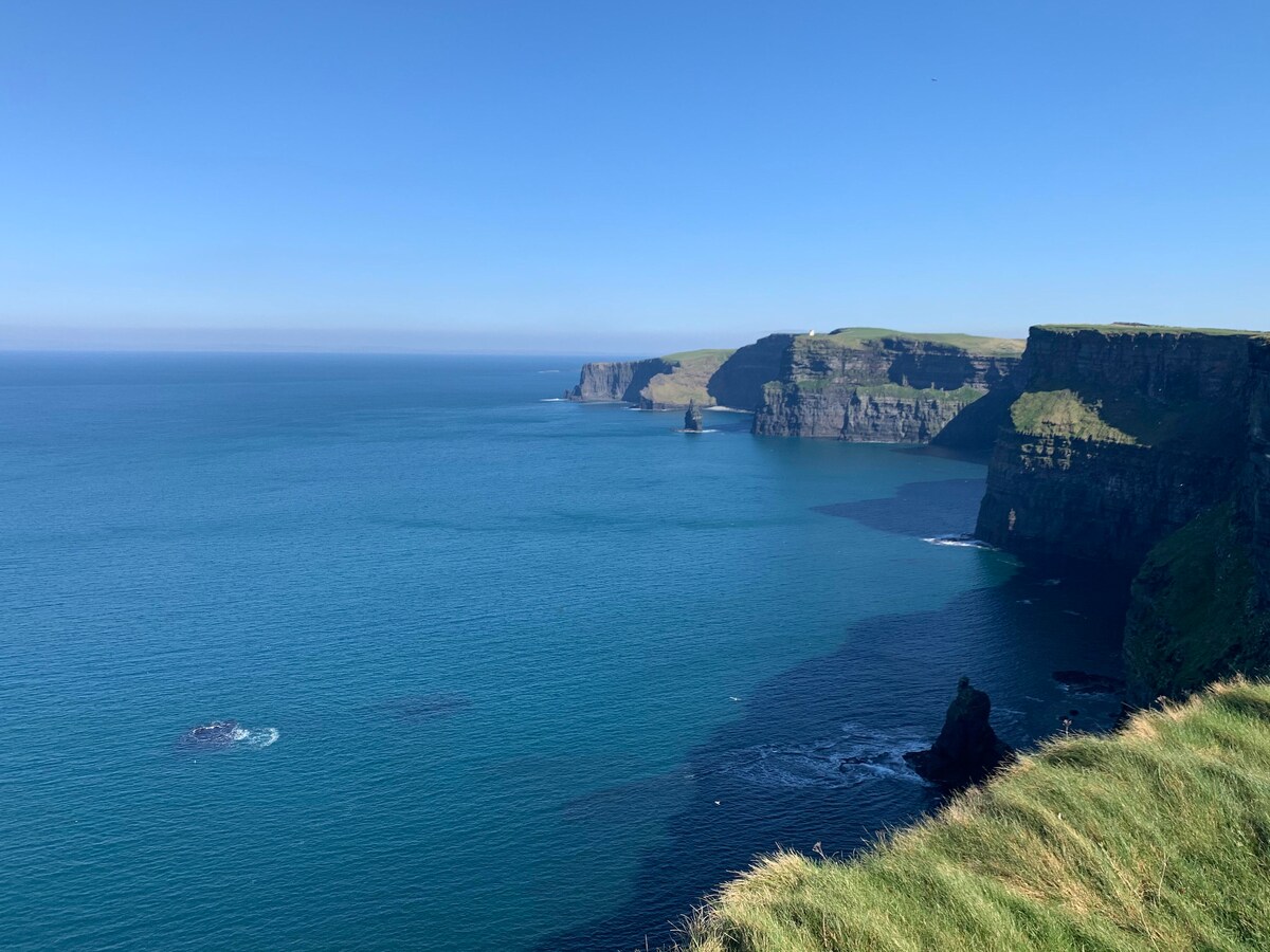 The image captures a panoramic view of the Cliffs of Moher, showcasing steep cliffs rising dramatically from the turquoise Atlantic Ocean. Gentle rolling hills line the horizon under a clear blue sky, creating a serene coastal landscape.