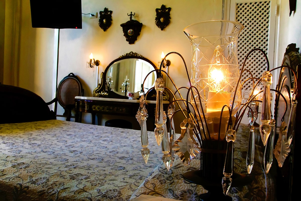 A spacious room features a large bed with intricate bedding, highlighted by a decorative lamp with crystal accents. A mirror and vanity can be seen in the background, complemented by vintage wall sconces on either side. Elegant details enhance the overall ambience.