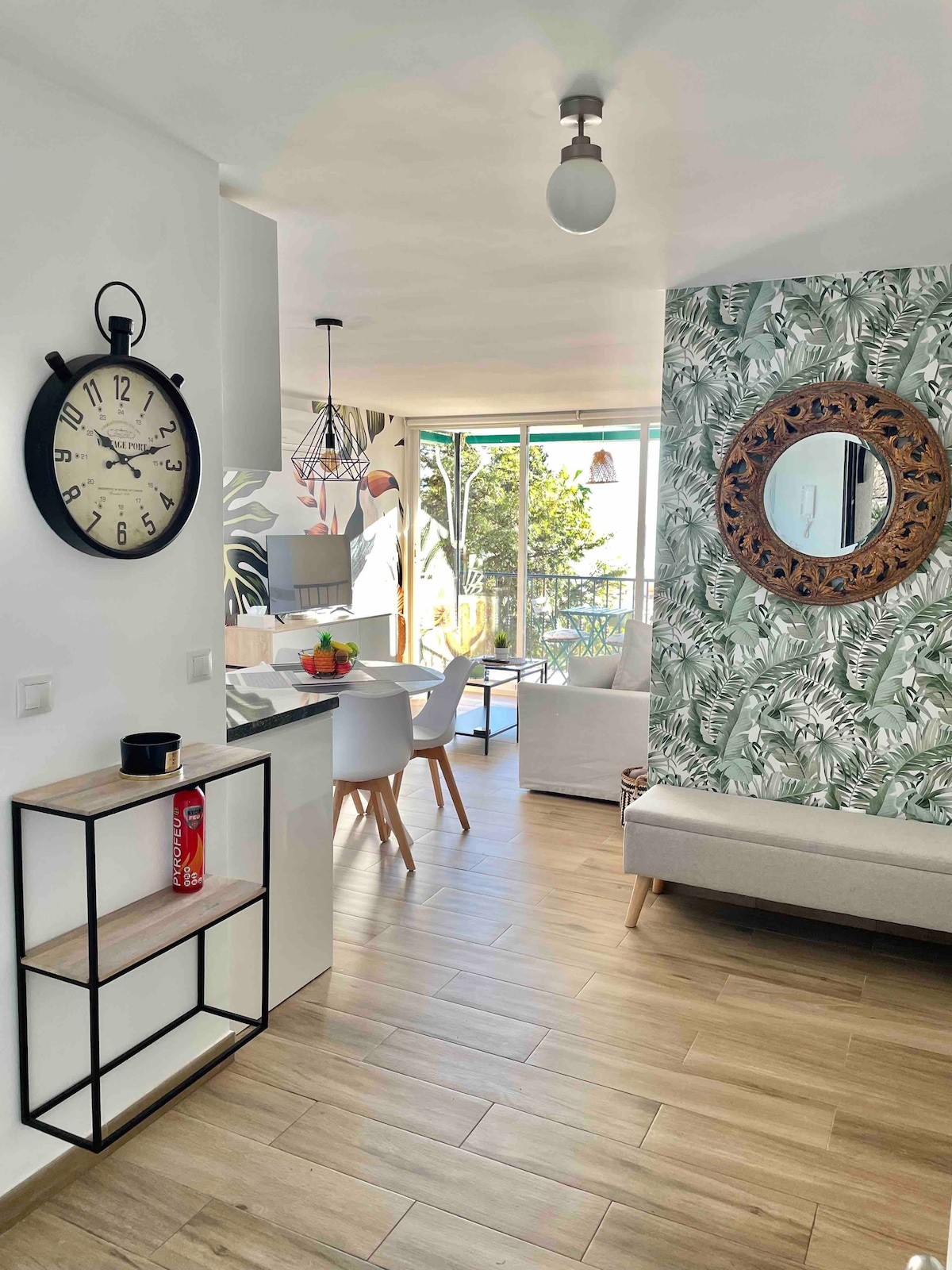 A modern living space features natural light flowing through large glass doors. A cozy sofa is positioned against a patterned wall adorned with tropical leaves. A round dining table with four chairs is visible, complemented by decorative accents including a wall clock and shelving unit.
