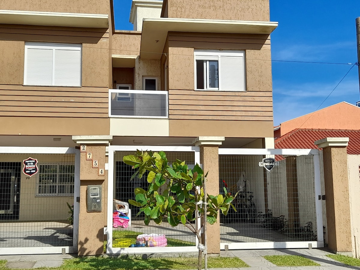 The duplex features a modern façade in earthy tones, highlighted by large windows that allow natural light. A well-maintained front yard includes a small tree and grass. The entrance is framed by a protective gate, ensuring security while allowing easy access.