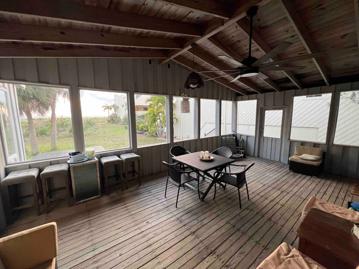 A spacious screened patio is featured, with wooden flooring and high ceilings. Seating options include a dining table and chairs, alongside casual lounge seating. Large windows provide views of the surrounding greenery and allow natural light to fill the area.
