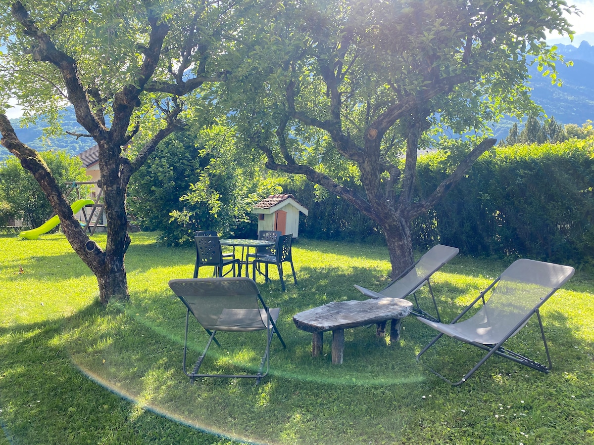 A garden area is displayed featuring two trees providing shade. A round table with four chairs is situated alongside two lounge chairs and a small wooden coffee table. A playhouse is visible in the background, surrounded by well-maintained grass.