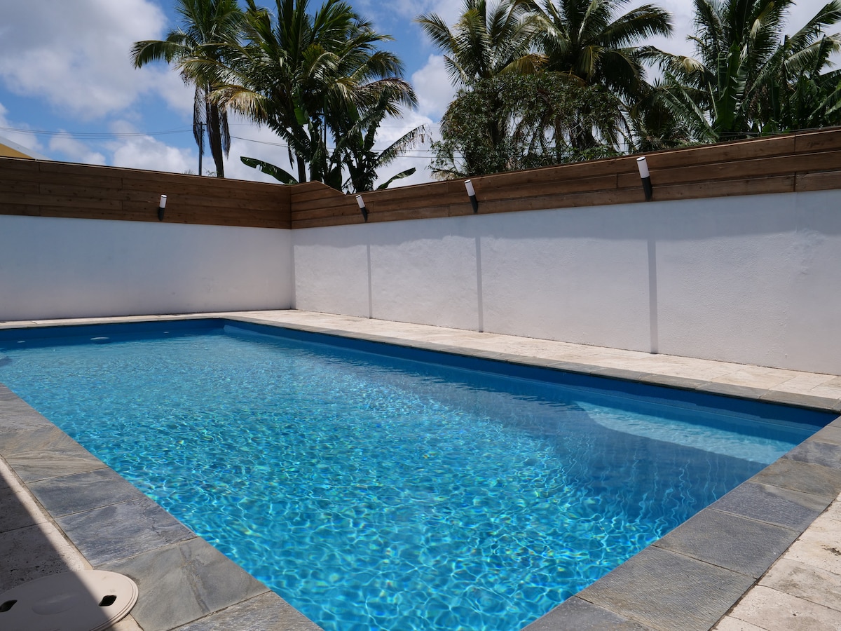 An exclusive swimming pool is situated within a private area, featuring clear blue water surrounded by tiled decking and palm trees. The walls are adorned with wood accents, providing a serene atmosphere. Natural light enhances the inviting pool area.
