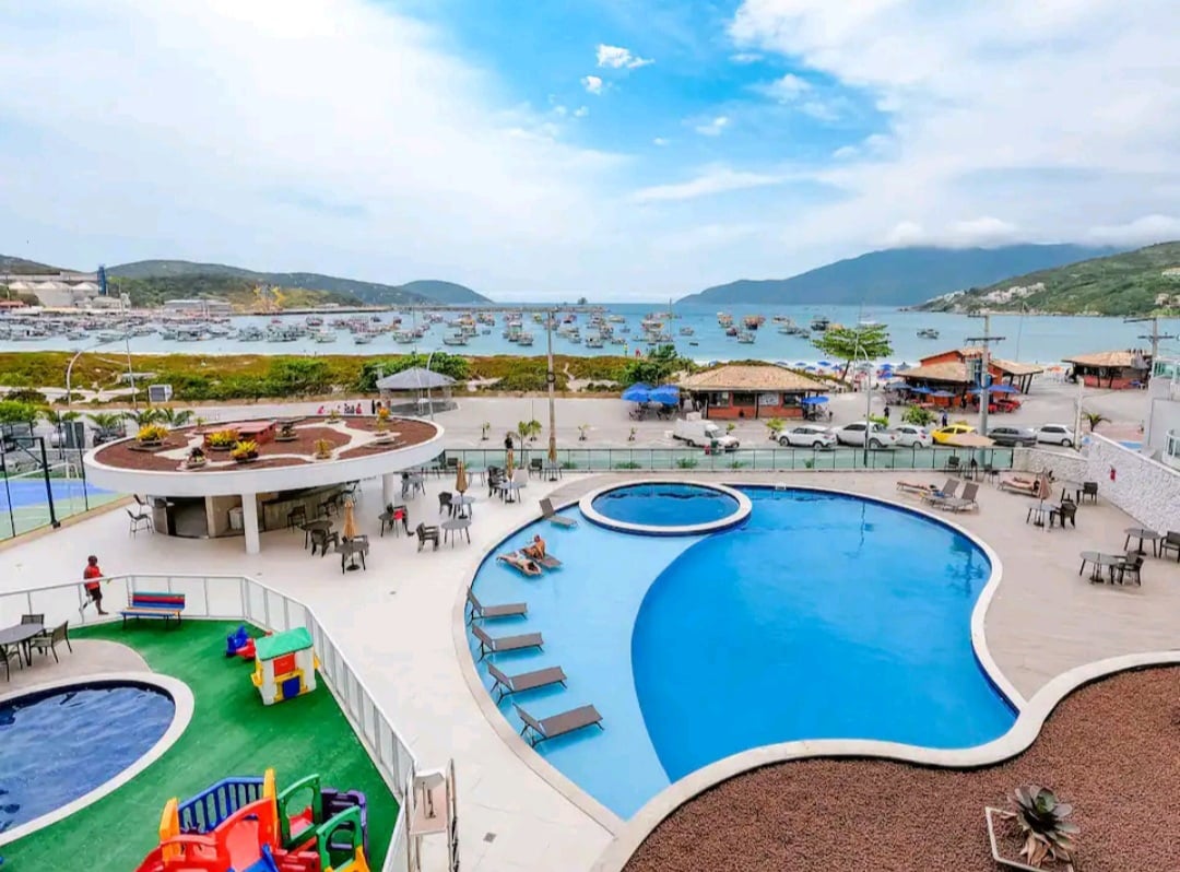 An expansive outdoor area features two swimming pools surrounded by loungers, with views of the marina and mountains in the background. A children's play area is visible, and several shaded seating arrangements can be seen throughout the space, offering diverse leisure options.