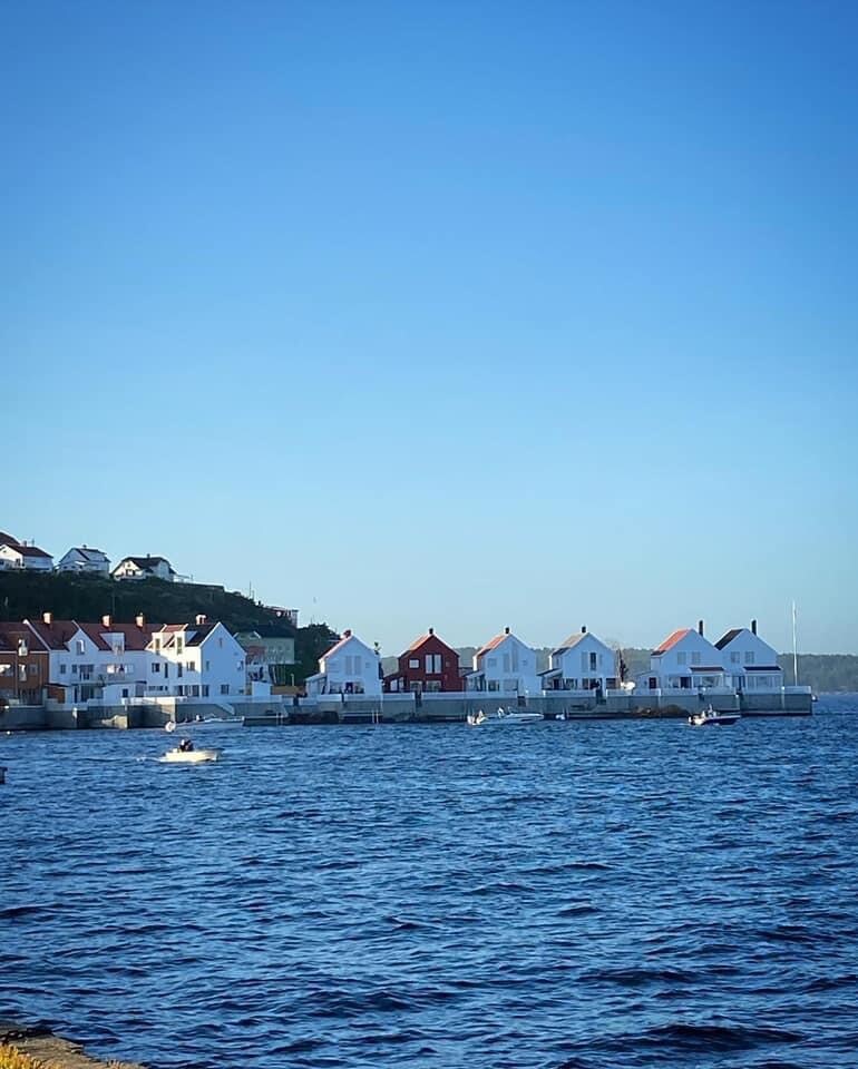 A row of charming houses, primarily in white with some red accents, stands along the waterfront. The calm blue water reflects the clear sky, while hills are visible in the background, contributing to the serene coastal setting.