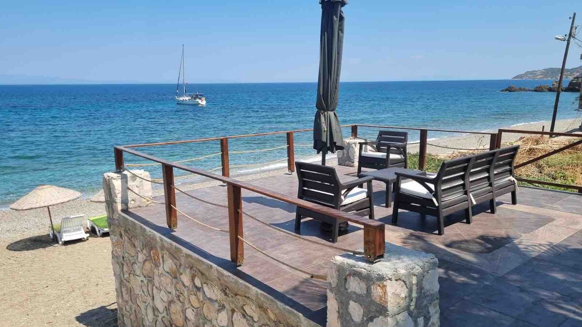 A spacious terrace features wooden seating arrangements, offering a panoramic view of the sea. The area is surrounded by a stone wall, with an umbrella providing shade. The calm waters and distant sailboat create a serene backdrop for relaxation.