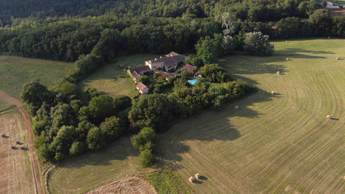 An aerial view captures an expansive property nestled within lush greenery. The main building is surrounded by trees and open fields, with a tranquil pool visible amid the landscape. Hay bales are scattered across golden fields, adding texture to the serene countryside scene.